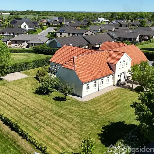 Et hvidt hus med rødt tegltag står stille i en velplejet have omgivet af grønne træer og hække. I baggrunden ses en velordnet villakvarter med flere huse.