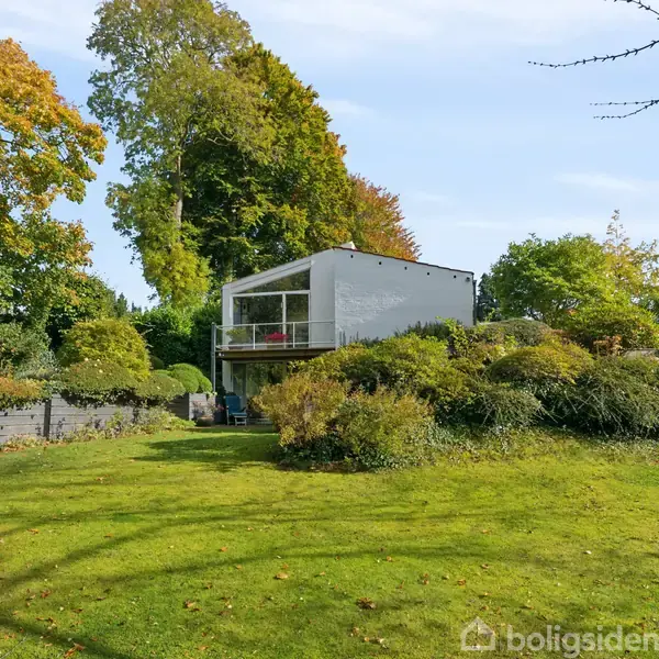 Et moderne hus står stille på en velplejet græsplæne omgivet af frodig beplantning og høje træer i efterårsfarver. En åben veranda er synlig på husets første sal.