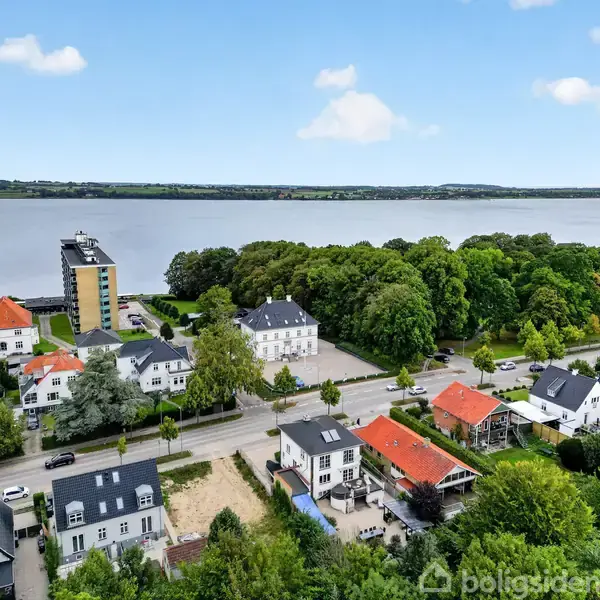 Flere huse står langs en vej nær en bred flod. Bygningerne har forskellige farver og former, omgivet af grønne træer. I baggrunden ses en højere bygning tæt på vandet.