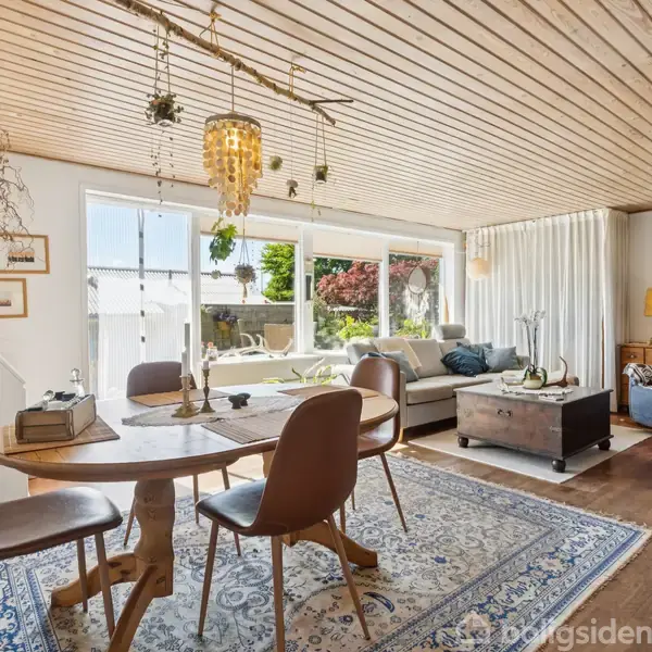 Et elegant opholdsområde indeholder en rund træspisebord med stole, sofaer ved store vinduer, rige på naturligt lys, og dekoreret med planter og kunstværker på væggene i en hyggelig stue.