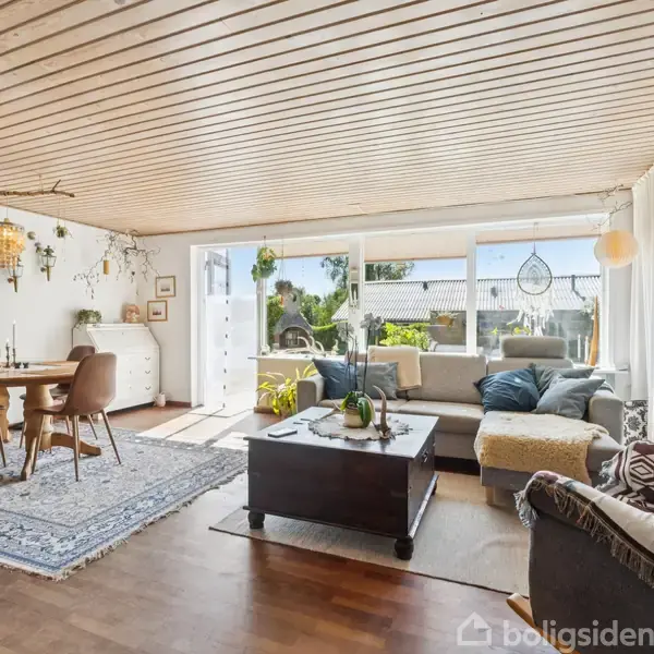 Stue er indrettet med spisebord og stole på et tæppe, sofagruppe med bord, store vinduer mod en terrasse med planter. Loftet er i lyst træ.