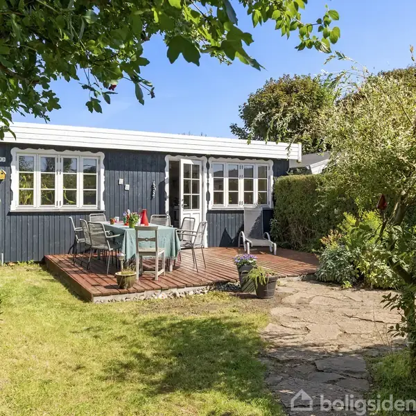 Et lille, sortmalet sommerhus med hvide vinduer står i en frodig have. På den træbelagte terrasse er et bord og stole placeret. Solen skinner, og træer omkranser området.
