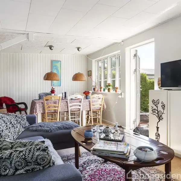 Stue med en blå sofa og et træbord fyldt med bøger og små genstande. I baggrunden ses spisebord med stole og to lamper over. TV står ved siden af store vinduer med udsigt.