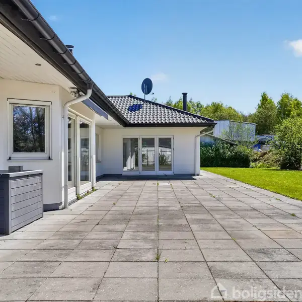 Huset har en stor flisebelagt terrasse med hvid facade og mørkt tag. I baggrunden ses en stor have med græs og træer under en klar himmel.