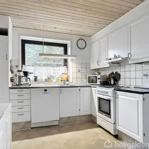 Hvidt køkken med komfur og opvaskemaskine står langs væggen. Omgivet af overskabe, fliser på væggen og et vindue med udsigt til træer. Vægur hænger over skabene.