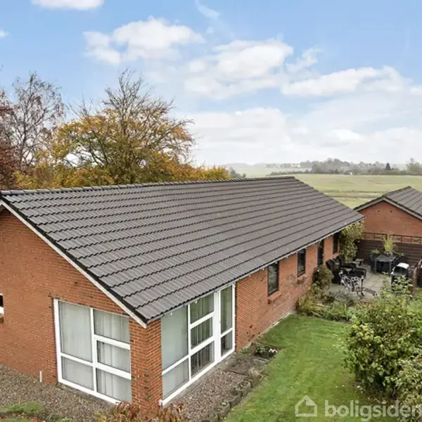 Et rødt murstenshus med gråt tag står i en have med græs og buske. En mindre bygning ses i baggrunden under en blå himmel med skyer.