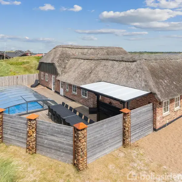 Stråtækt hus med indhegnet terrasse og overdækket pool. Beliggende i et sandet, åbent landskab med spredte huse i baggrunden og en klar himmel med lette skyer.