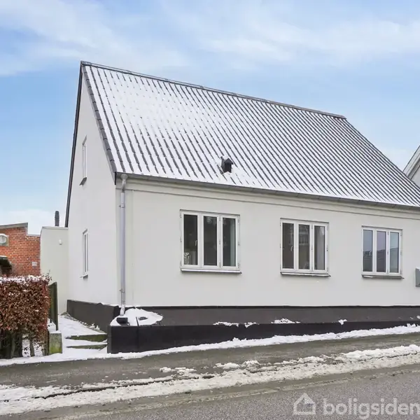 Et hvidt hus med skråt tag er dækket af sne. Huset står på en snebeklædt vej. Nummeret "33" er synligt på facaden. Ved siden af huset står en gadelampe.