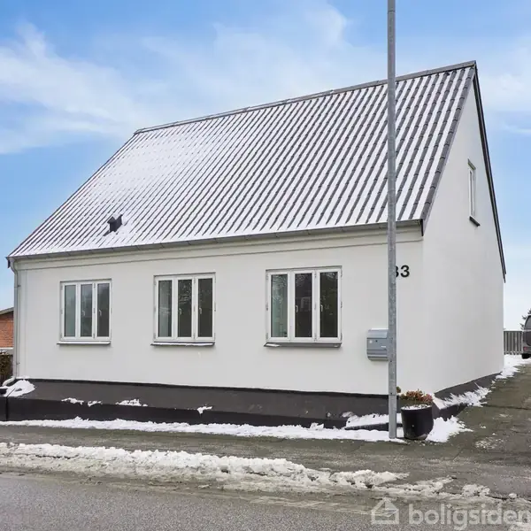 Et hvidt hus med skråt tag, beliggende ved en vej med lidt sne på jorden. Huset har fire vinduer og en gadelygte foran. Til venstre ses en høj hæk.