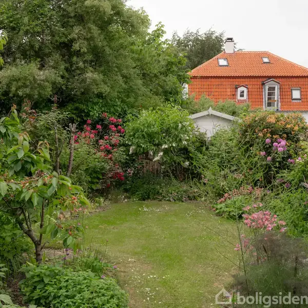 Et hus med orange tagsten er omgivet af en frodig have med forskellige blomstrende planter og træer. Stien fører gennem den grønne vegetation.