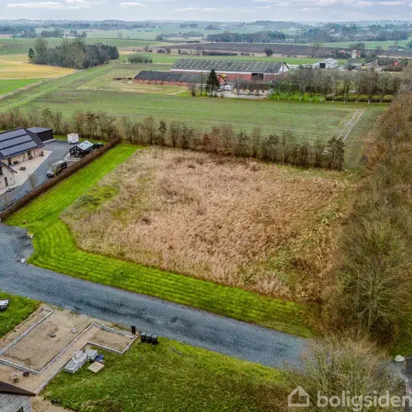En mark ved siden af en vej, hvor en bil passerer. I baggrunden ses huse, marker og et skovbryn.