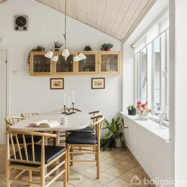 Et lyst køkkenalrum med et ovalt spisebord og fire stole. Bordet har en åben bog og lysestager. Et træloft med en lampe over bordet, og en væghylde med dekorationer.