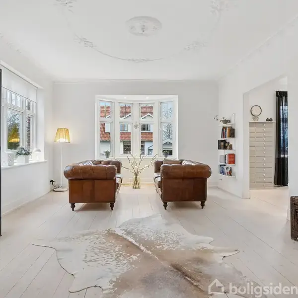 To lænestole står foran et stort vindue med udsigt til bygninger. Et glasvitrineskab og en standerlampe er til venstre. Et tæppe ligger på det lyse trægulv.