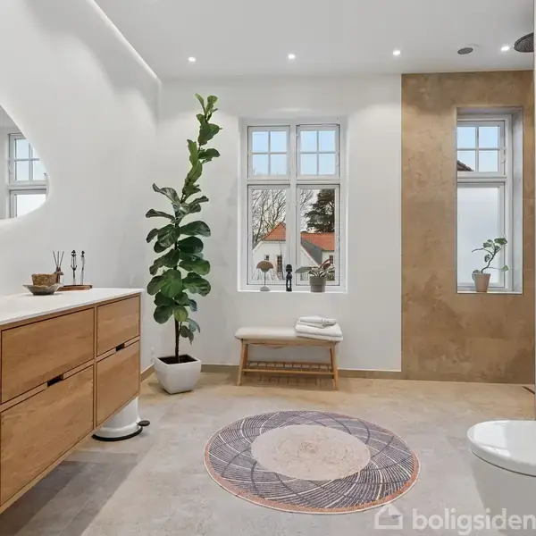 Badeværelse med træskabe står langs væggen under et stort spejl. Plante ved vindue, cirkulært tæppe på gulvet. Toilet til højre, kunstværk hænger på væggen. Ingen tekst fundet.