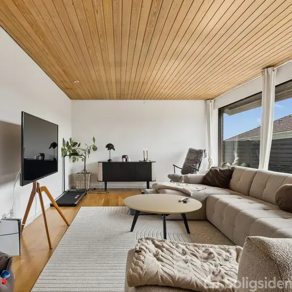 Stue med stor beige sofa, sofabord og fladskærm. Lysindfald fra store vinduer, træloft og planter som dekoration. Åben og indbydende atmosfære, moderne indretning.