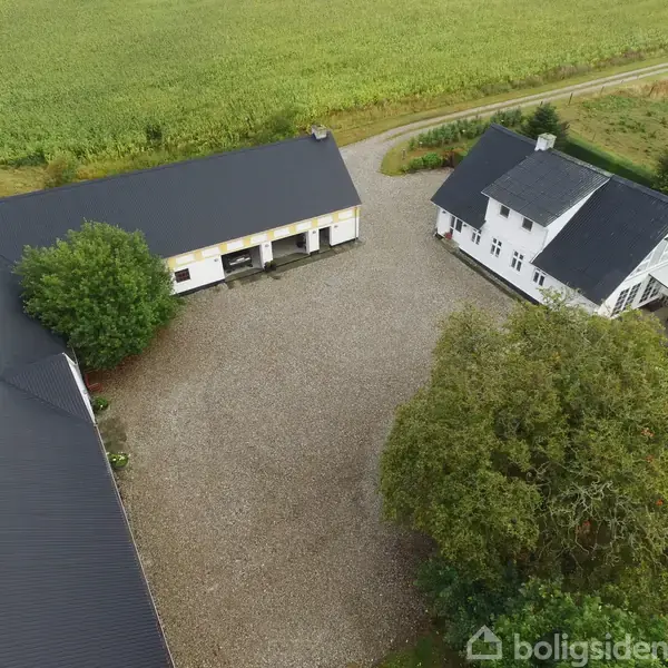 Stuehus og ladebygninger omkranser en gårdsplads med grusbelægning; træer og marker omkranser gården.