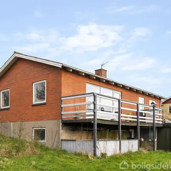 Et rødt murstenshus har en balkon med metalgelænder, omgivet af en grøn have og træt miljø. Huset har flere vinduer og en skrånende tagrytning under en klar himmel.