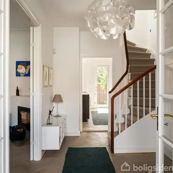 En indgangshal med en moderne lysekrone hænger fra loftet, trappen fører opad til anden etage. Rummet har hvide vægge, trædøre og et tæppe på gulvet.