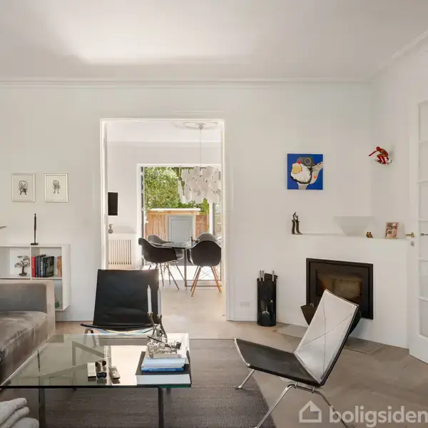 Stue med grå sofa og glasbord, åbner mod spisestue med sorte stole. Pejs ved siden af moderne lamper og vægdekoration. Lysindfaldet skaber en åben og hyggelig atmosfære.