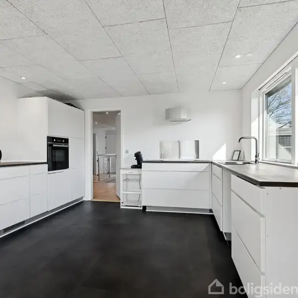 Et moderne køkken med hvide skabe og sorte bordplader. En elkedel står på køkkenbordet. Store vinduer giver udsigt til en have. En plakat hænger på væggen.