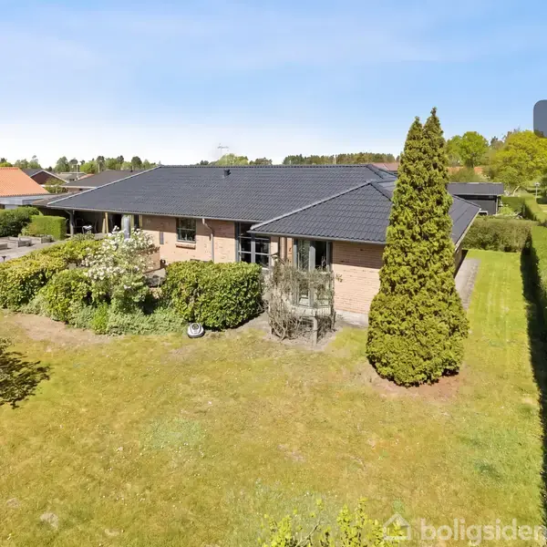 En murstensvilla med sort tag, omgivet af en velplejet have med buske og træer, placeret i et stille boligområde. Tekst: "Visualisering".