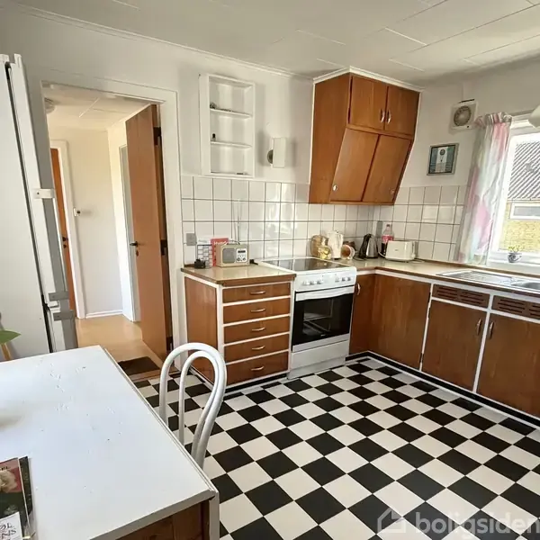 Et køkken med komfur, brune skabe og sort-hvidt ternet gulv. Et lille bord med stol står ved vinduet. Lyse vægge og fliser skaber en lys atmosfære.