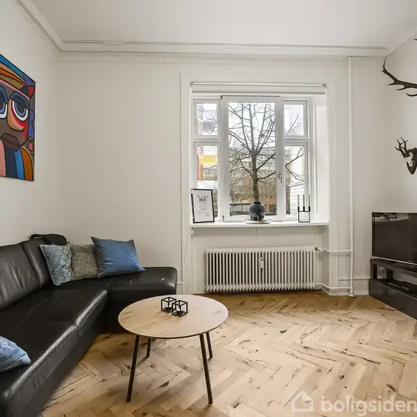 En sort lædersofa står i en stue ved siden af et rundt træbord. Væg med farverigt maleri. Vinduet viser et træ udenfor. Jagt trofæer hænger over et TV.