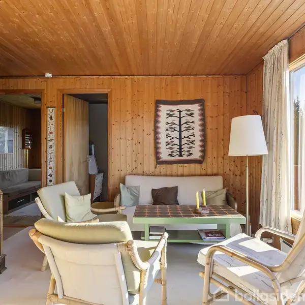 Stue med to lænestole og en sofa, omgivet af træpaneler og store vinduer. Et sofabord står centralt, dekoreret med et tæppe på væggen og en standerlampe i hjørnet.