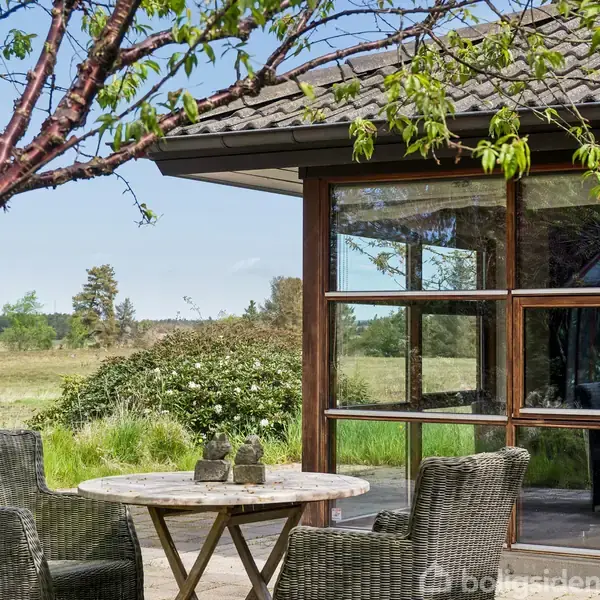 En terrasse med to flettede stole og et rundt bord står foran et glashus. Omgivet af en grøn natur med træer og buske i baggrunden.