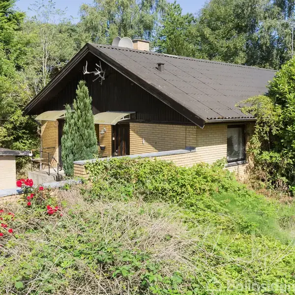 Et gult hus med brunt tag står omgivet af frodige buske og træer. En ståltrappe fører til hoveddøren. Der er en terrasse til venstre med røde blomster omkring.