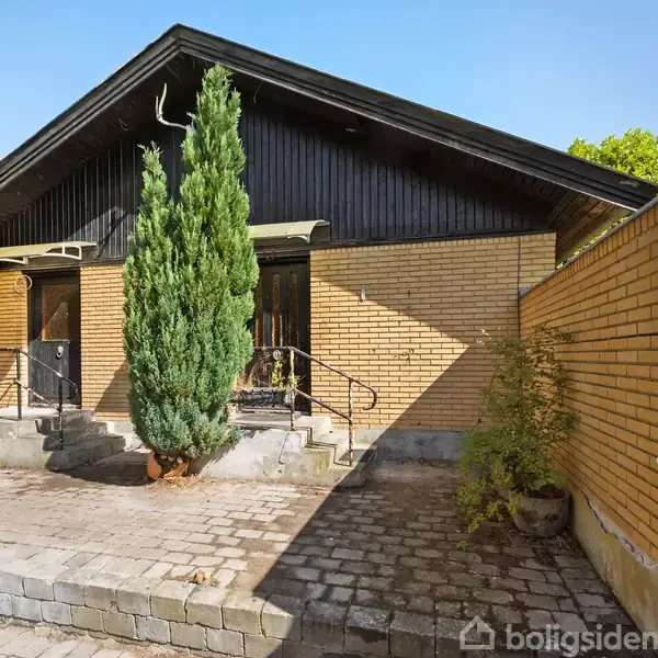 En gulstensbygning med sorte detaljer, centralelement er en stor grøn busk foran trappen. Omgivet af en flisebelagt gårdsplads og en tilstødende mur.