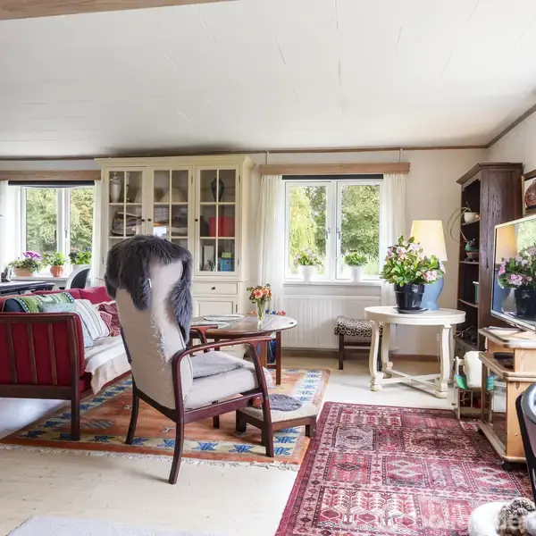 En lys stue med et komfortabelt siddemøblement i form af en lænestol og sofa. Der står et fjernsyn på en stander, og rummet er omgivet af store vinduer med udsigt til grøn natur.