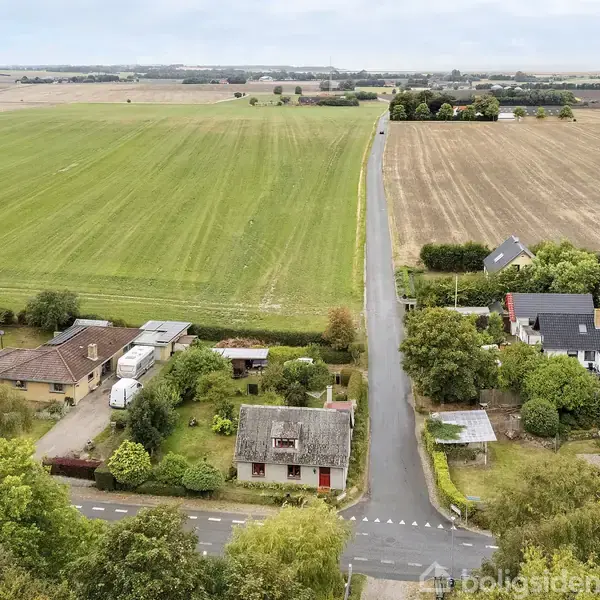En lille landsby med huse spredt omkring en vej, omgivet af vidtstrakte marker og grønne træer. Hovedvejen strækker sig langt ud i landskabet med bygninger på begge sider.