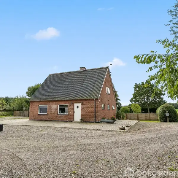 Et rødt murstenshus med gråt tag står på en grusbelagt gårdsplads. Omkring huset er der grønne træer og marker under en blå himmel.