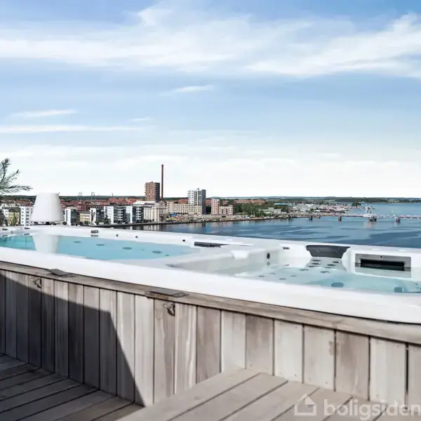 Udendørs spa står på en træterrasse med udsigt over en by beliggende ved vandet. I horisonten ses bygninger og skorstene under en blå himmel.
