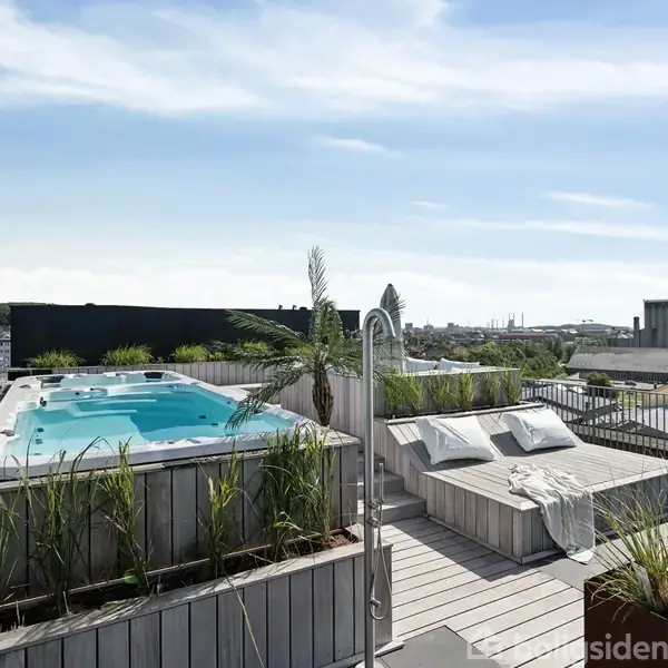 En jacuzzi står på en tagterrasse omgivet af planter og liggestole med hvide puder, med udsigt over en by med høje bygninger i horisonten under en blå himmel.