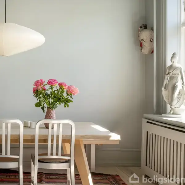 En vase med lyserøde blomster står på et lyst træbord i et minimalistisk rum. To stole er ved bordet, og en statue samt en maske er placeret ved vinduet.