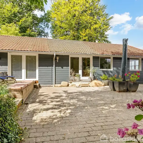 Blå-gråt træhus med rødt tag står i solskin; store vinduer åbner mod en flisebelagt terrasse med planter i krukker. Omgivelserne inkluderer blomsterbede og frodig beplantning.