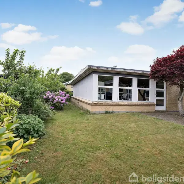 En murstensvilla med store vinduer står stille i en grønn have med farverige blomsterbuske og et rødt træ. Skyfri himmel med skyer i baggrunden.