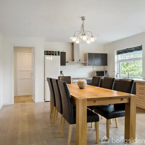 Spisestue, bord med otte sorte stole, står i rummeligt, lyst køkken. Moderne køkkenelementer og vinduesudsigt. Lampe hænger over bordet, træ- og hvidfarvet indretning.