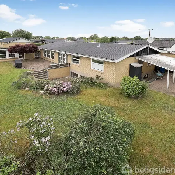 Et enfamiliehus med fladt tag står omgivet af en grøn have med buske og et overdækket område til højre. Baggrunden viser en række af træer og flere huse i det fjerne.