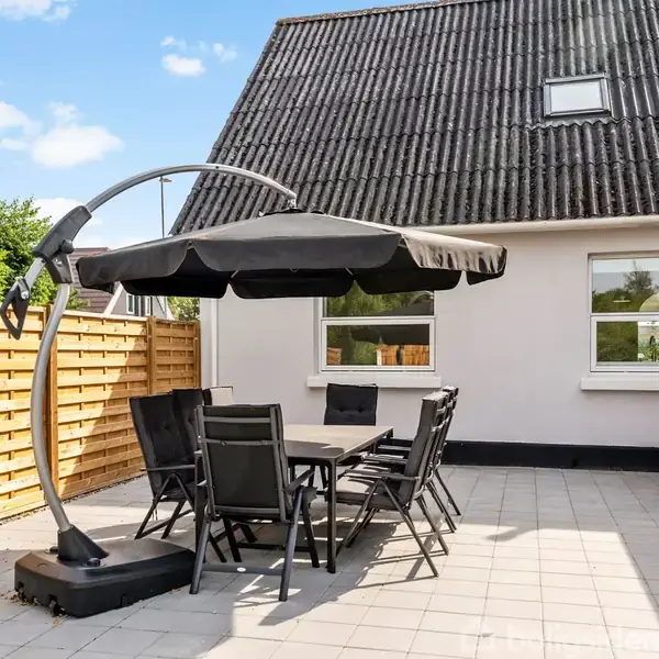 Et havebord med seks stole under en stor, rund parasol står på en flisebelagt terrasse. Baggrunden er et hus med gråt tag og hvide vægge, omgivet af et trægærde.