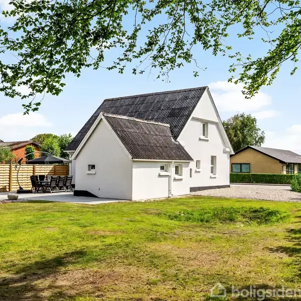 Et hvidt hus med sorte tagsten står i en have omgivet af grønne træer og græsplæne. Der er en terrasse med havemøbler til venstre.