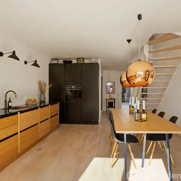 Et moderne køkken med træskabe og sort bordplade, hvor knive hænger på væggen. En lang spisebord med seks stole står i midten. Trappe fører opad.