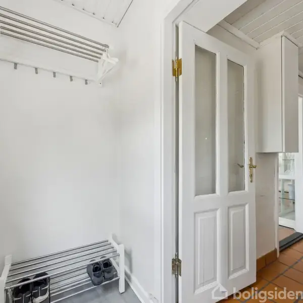 En entre med en skohylde og en knagerække i venstre side. En hvid dør med glasruder åbner ind til et køkken med flisegulv og hvide skabe.