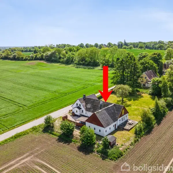 Hus markeret med rød pil, står på landet omgivet af grønne marker og træer med en vej i nærheden.