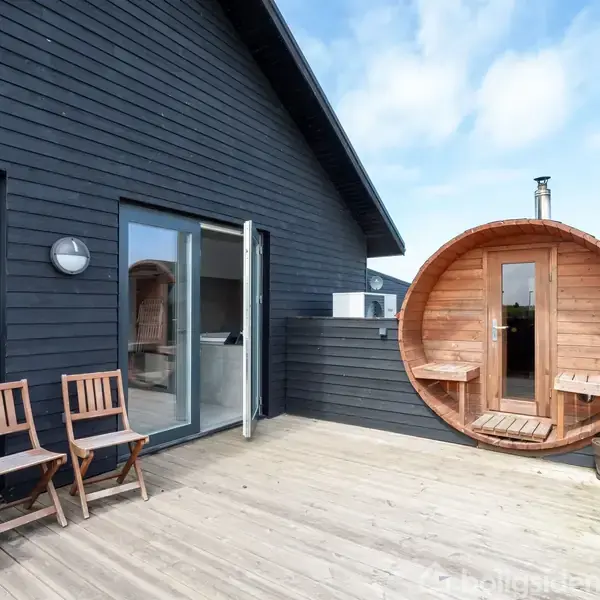 Et rundt, træsauna står stille på en stor, træterrasse. To træstole står ved siden af en glasdør. Bygningen er sortmalet med store vinduer i moderne stil.