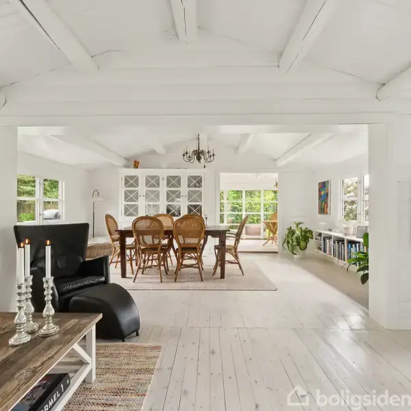 Stue med lyse trægulve og hvidt loft. Et bord med stearinlys står foran en lænestol. I baggrunden ses et spisebord med stole. Store vinduer tilføjer lys.