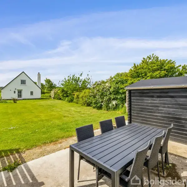 Et sort udendørs spisebord med stole står på en terrasse. I baggrunden ses en stor grøn græsplæne og et hvidt hus omgivet af buske og træer under en klar blå himmel.