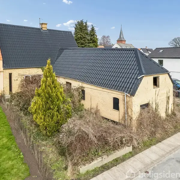 En gul murstensbygning med sort tegltag står omgivet af en grusvej og grønne omgivelser. En flagstang er placeret tæt på huset, og andre huse er synlige i baggrunden.
